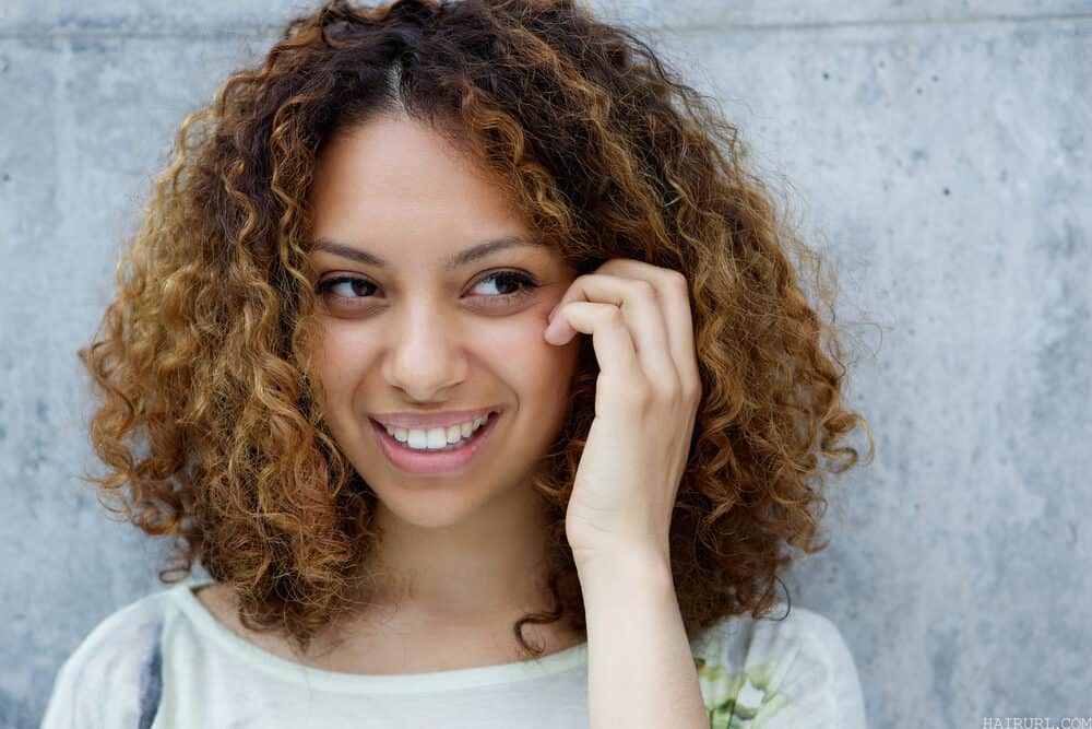 Light-skinned black girl with curly bleached hair wearing a big smile while touching her face