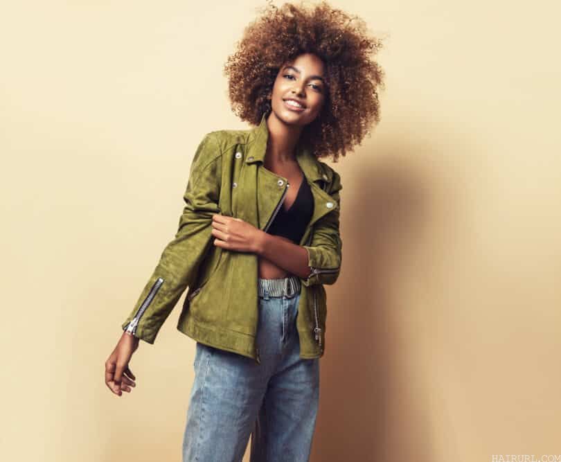 African American woman smiling while wearing a green winter jacket, blue pants, and hair that is naturally curly.
