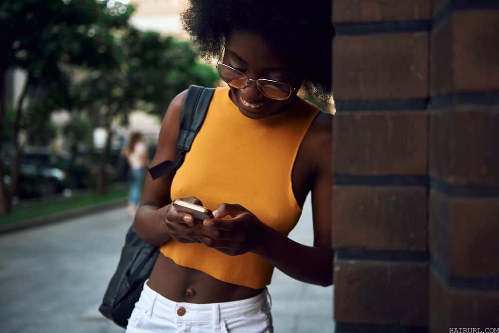 Woman with black natural hair wearing glasses and standing next to a brick wall while talking on her phone.
