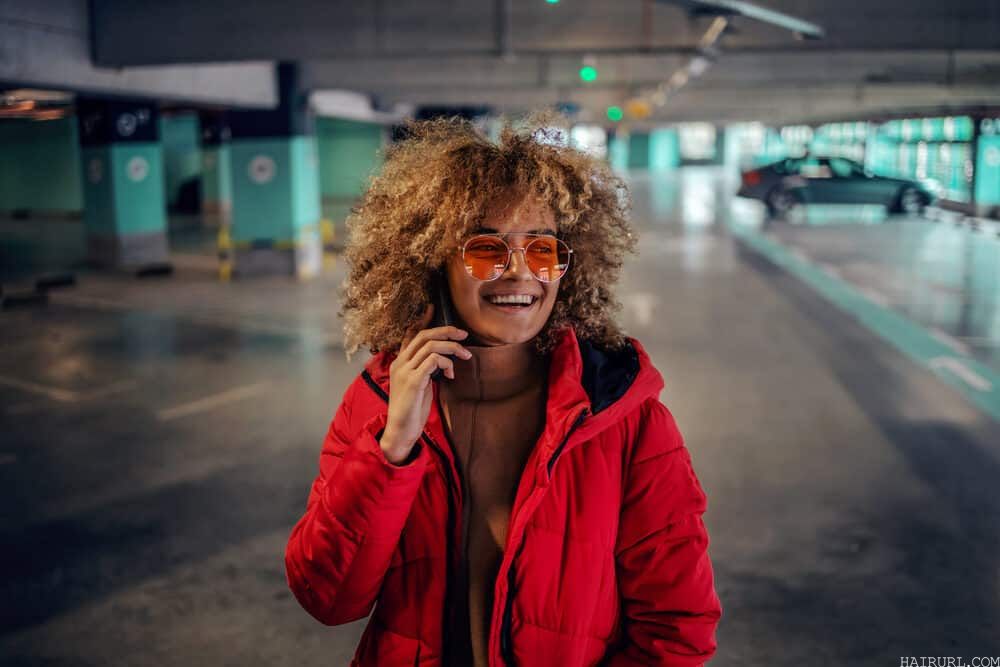 A young woman with curly hair, carrying a phone to her ear as she stands in the underground garage.