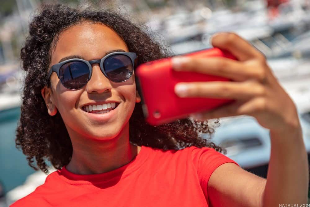 Beautiful African American woman  wearing sunglasses, a red t-shirt, and smiling with perfect teeth taking selfie photograph with red i-phone in the summer sunshine by a luxury boat filled harbor. The woman has very curly black and brown hair that's been moisturized with coconut oil.
