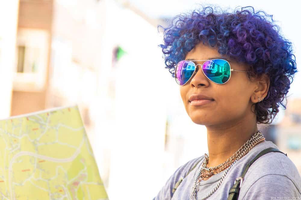 Female wearing rainbow shades, purple-blue hair, earrings, and natural lipstick color.