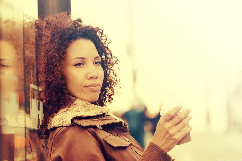 Black girl with naturally curly hair wearing a brown coat and drinking tea.