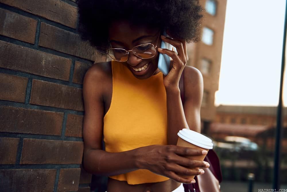 A smiling lady standing near a brick wall with a coffee while talking on her phone.
