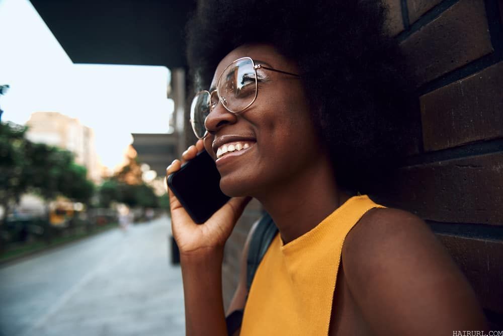 Cute black woman with naturally curly hair in glasses talking on the phone while leaning against a brick wall.