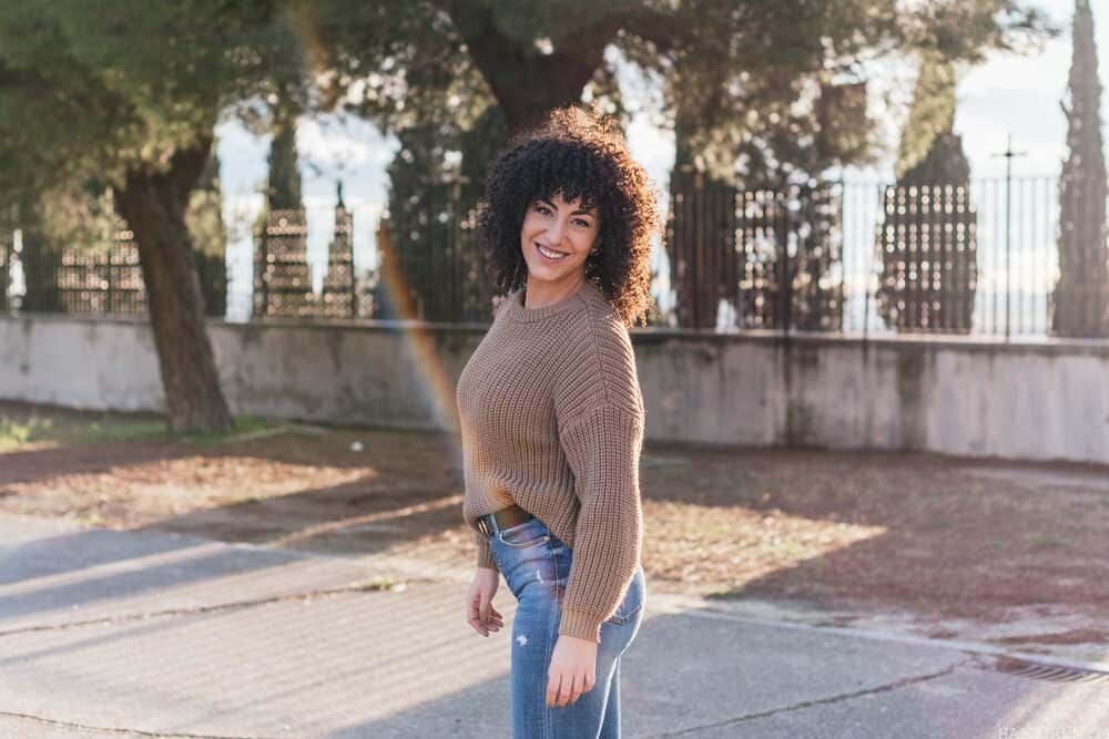 Pretty white adult woman with natural hair standing colored with expired hair developer in the park taking photos wearing a sweater, Gucci belt, jeans, and sneakers.