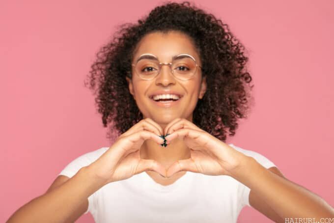 Female wearing a white t-shirt, eye shadow, and black fingernail polish, making a heart symbol with her hands.