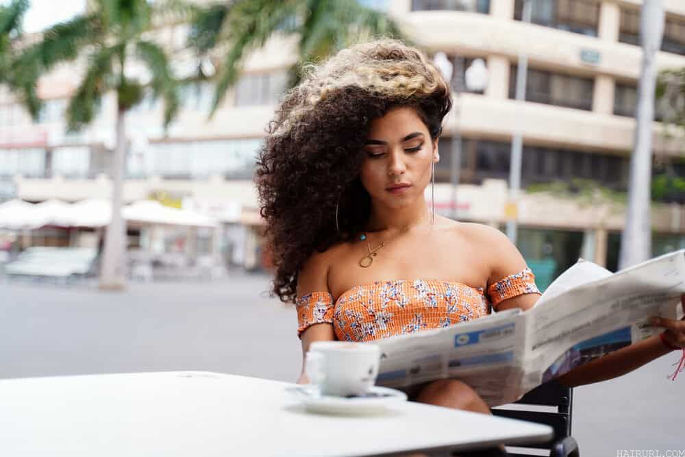 Beautiful woman reading a newspaper, while wearing hoop earrings and drinking a smooth liquid drink.