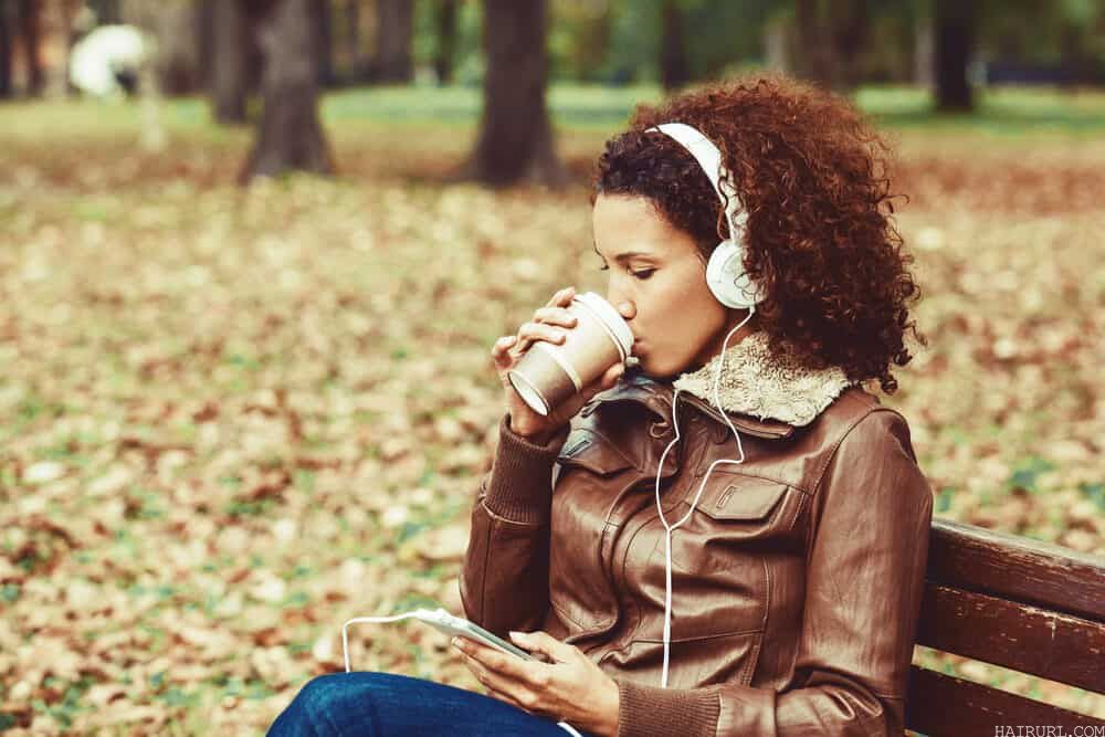 Women with 3c natural hair sitting in the park wearing a brown leather coat and drinking coffee.